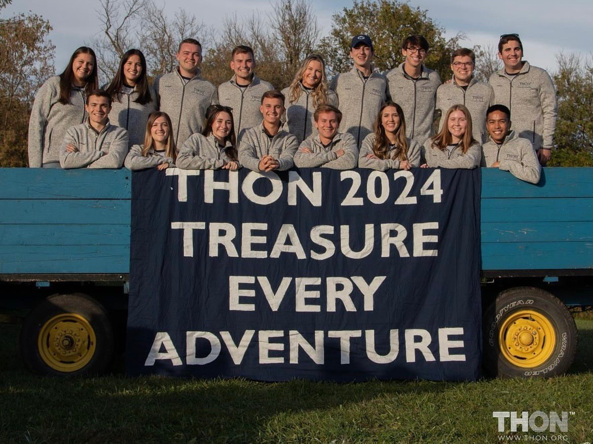 The THON 2024 directors stand in two lines on a trailer bed behind a banner that says, “THON 2024 Treasure Every Adventure.”