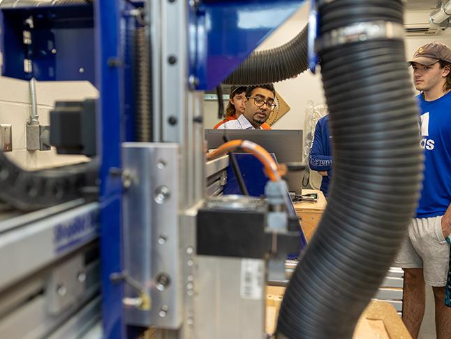 A professor stands among lab equipment while a student watches.