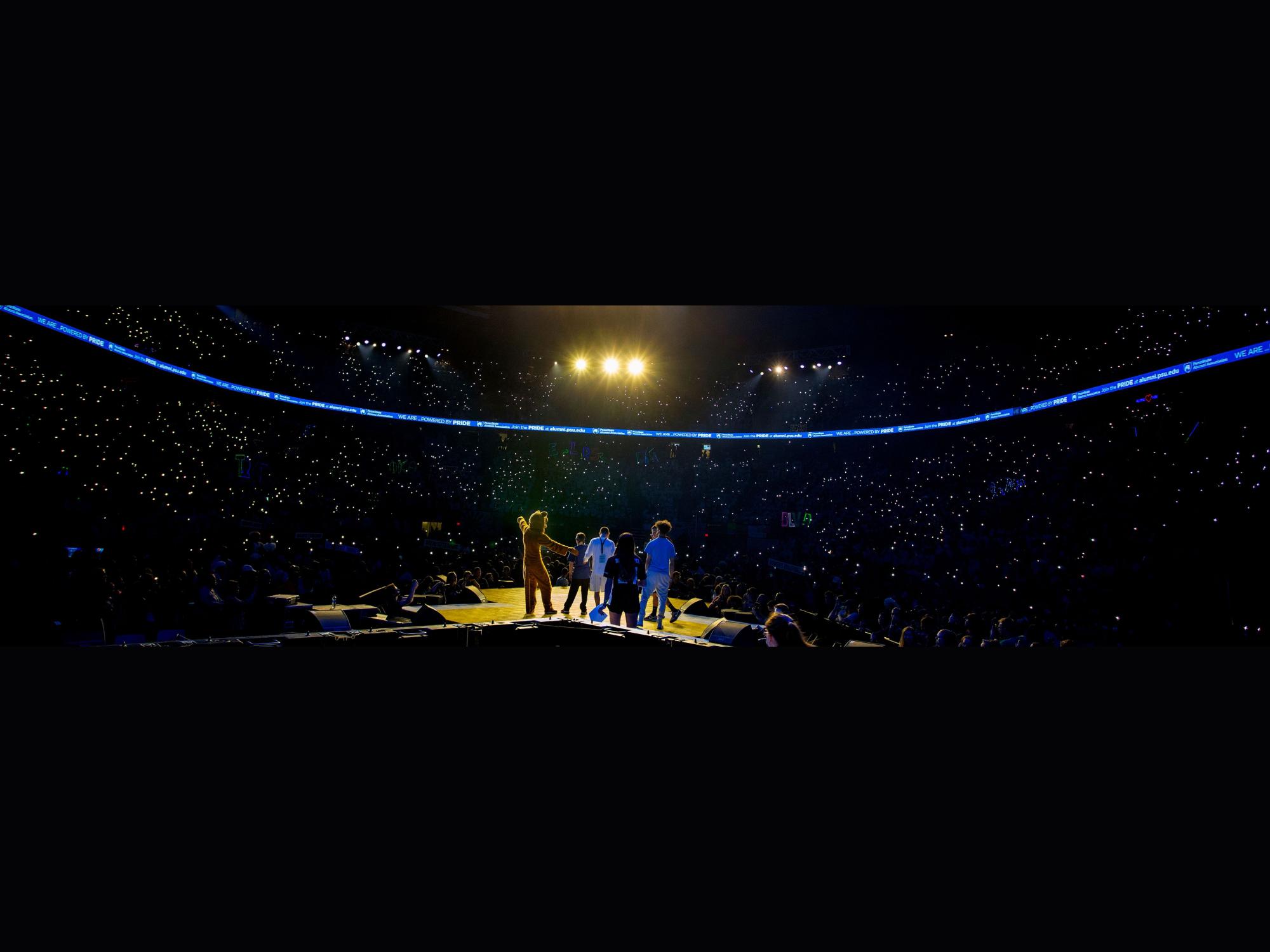 A rear shot of the Nittany Lion and others on stage facing the crowd in a dark arena