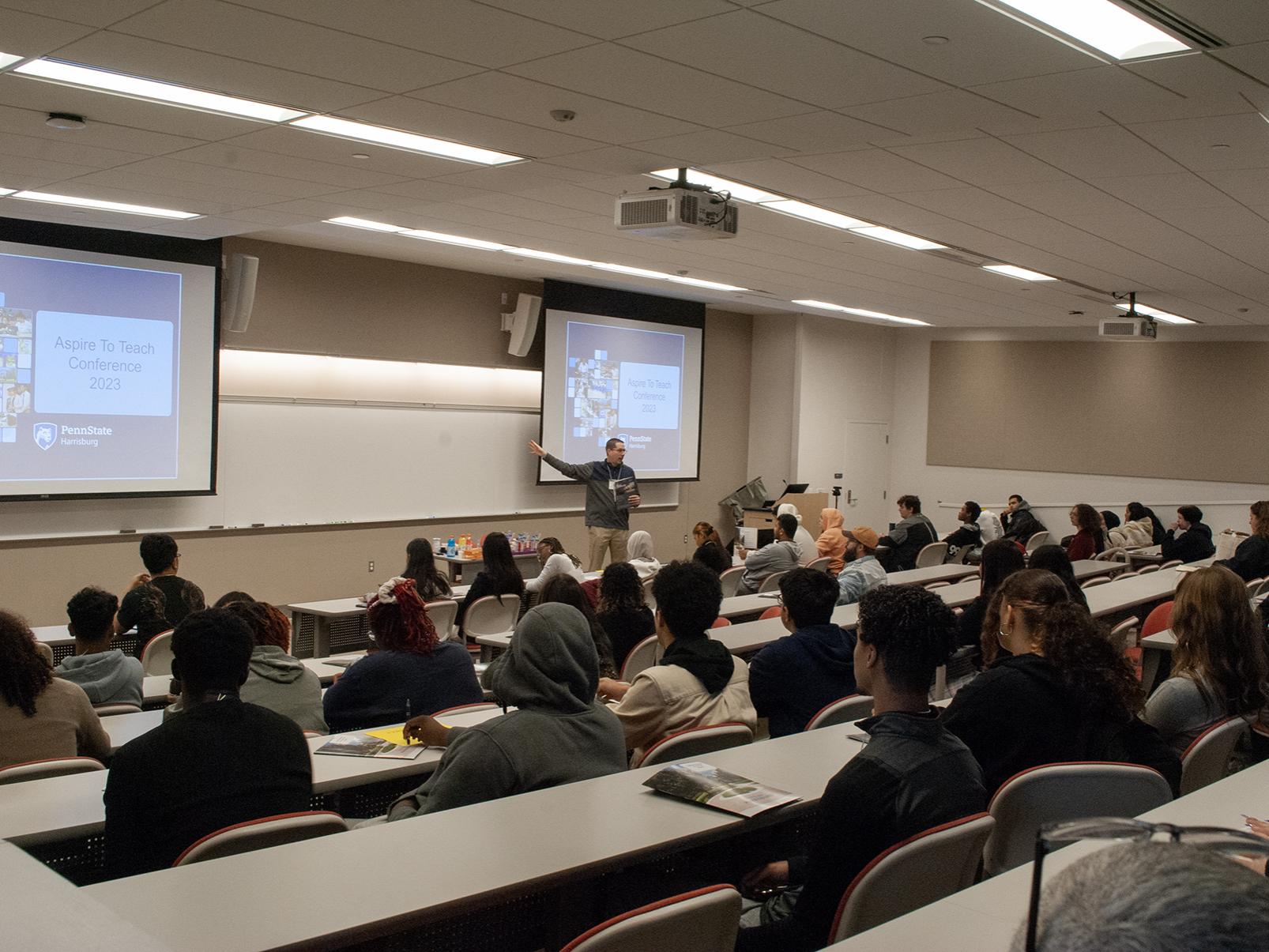High school students sit in a large classroom, where screens show the words Aspire 2 Teach Conference