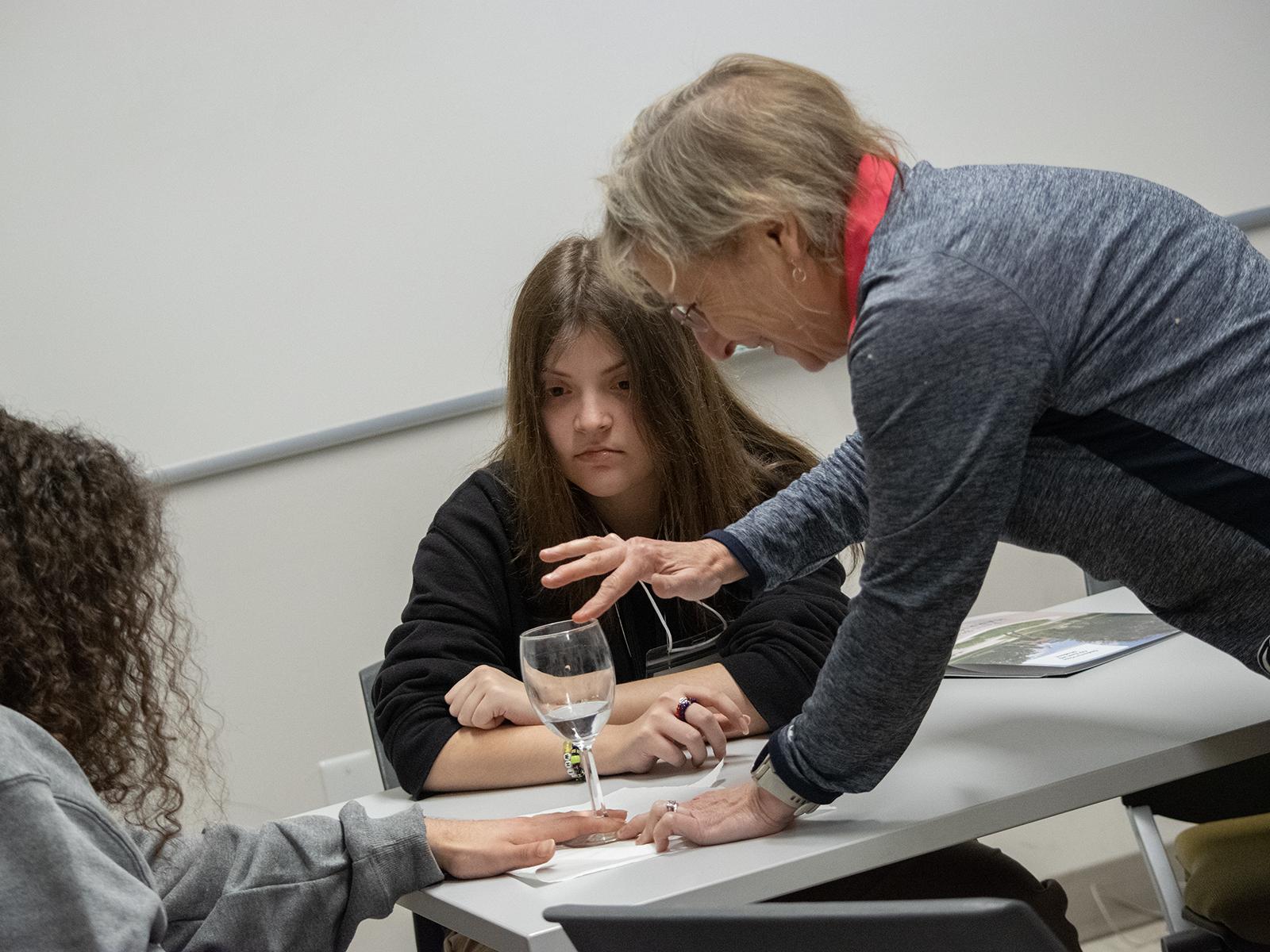 Students watch as a professor runs her finger on the rim of a glass containing water during a STEM activity