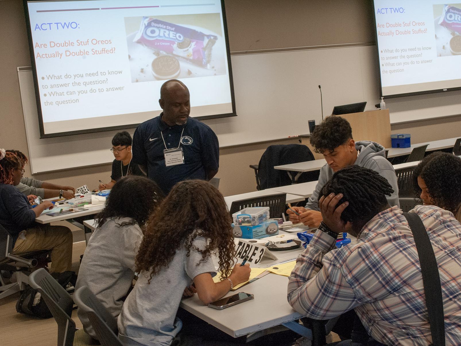 A professor overlooks a table, where high school students are working with scales and Oreos
