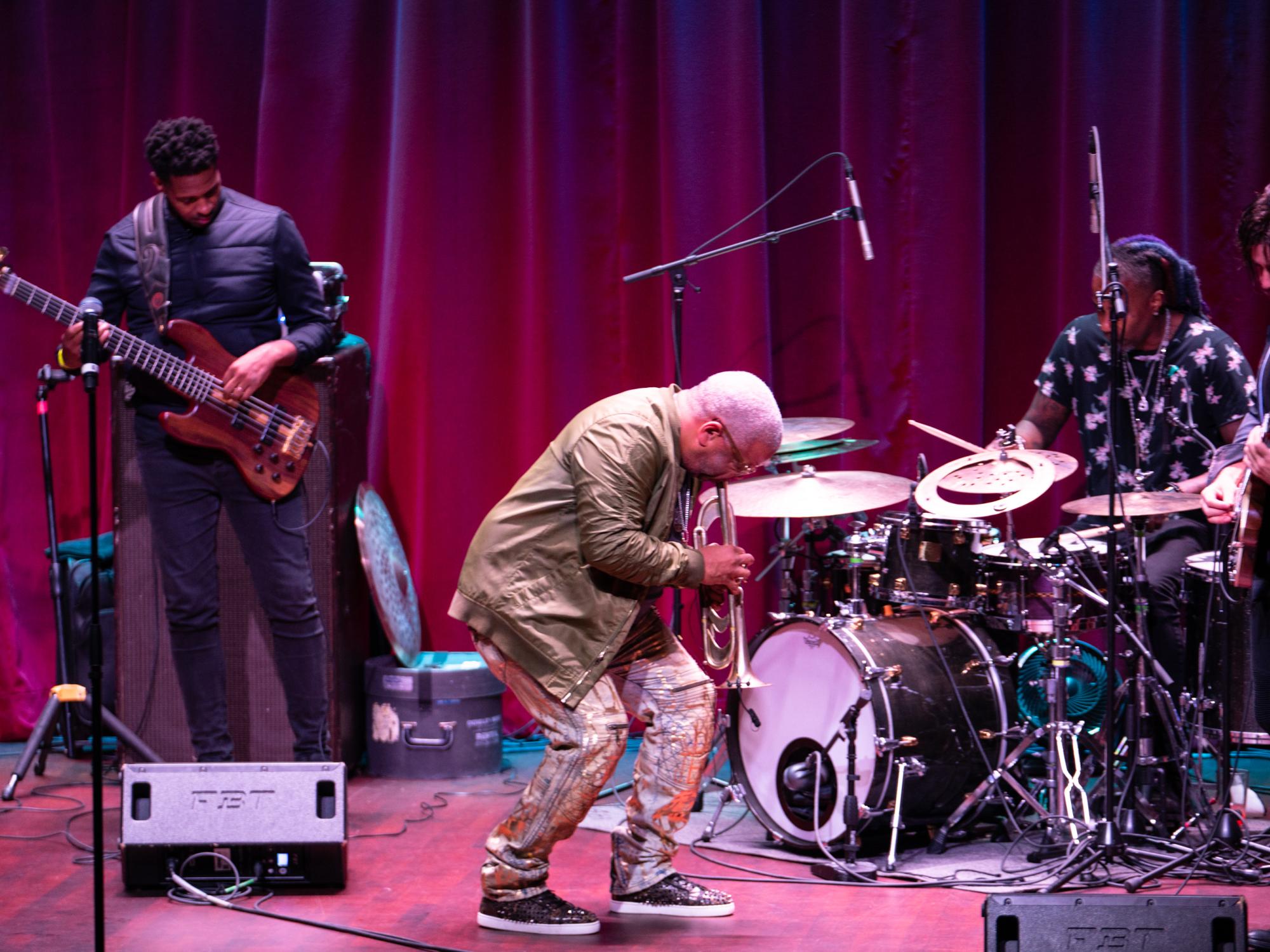 A man crouches down and plays a trumpet while surrounded by various jazz musicians.