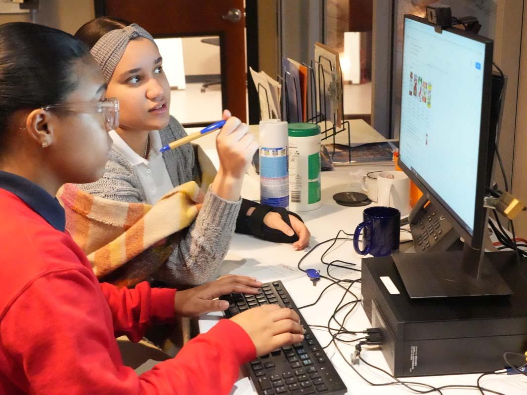 Two female students with pens and notebooks looking at a computer screen while seated at a desk.