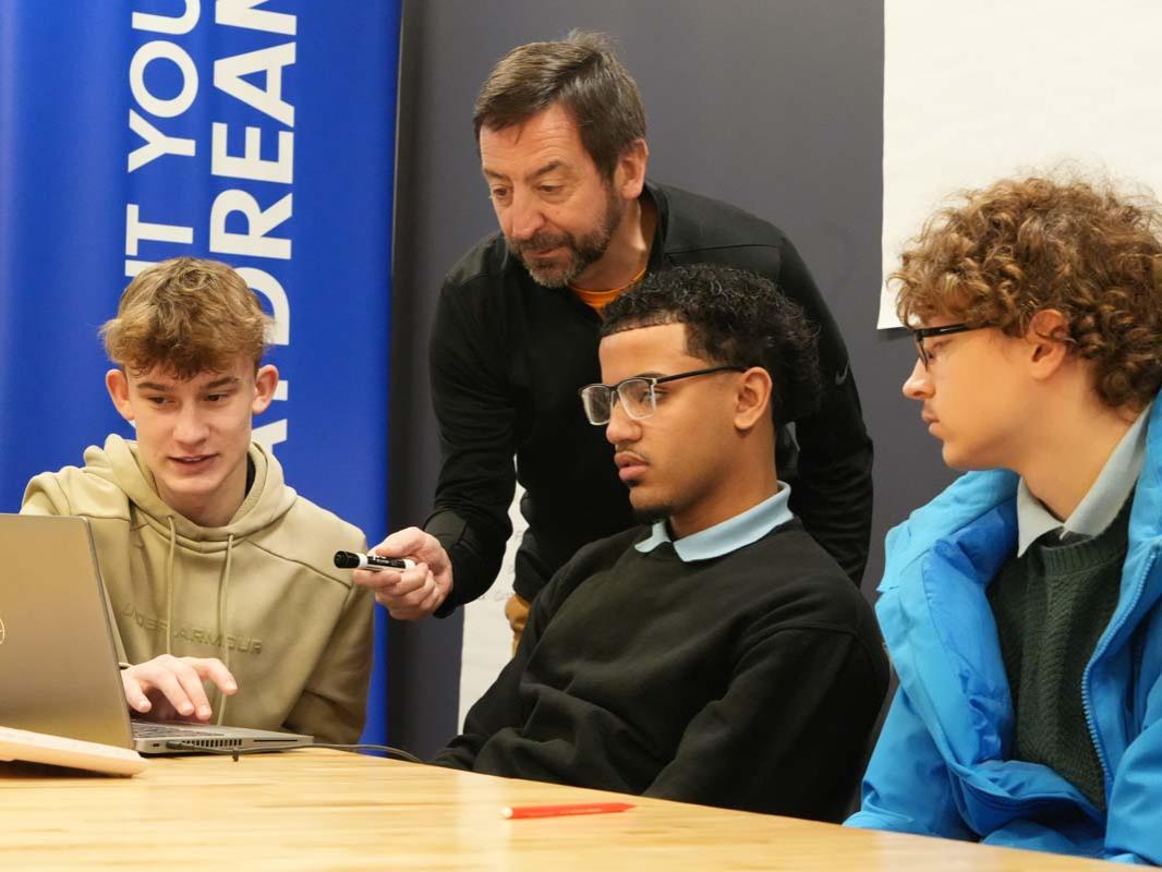 An instructor leaning over a table where three male students are working on a laptop.