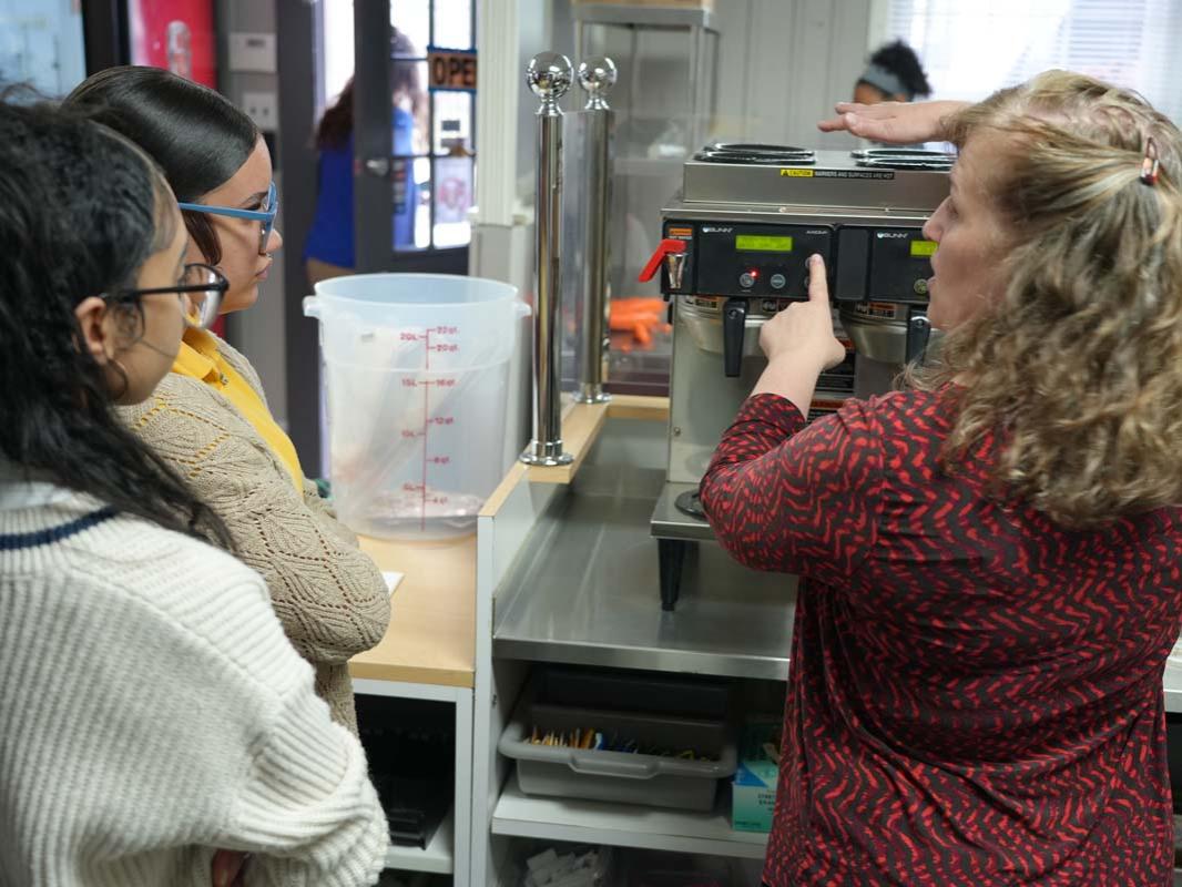 Woman holding coffee pot next to coffee machine while students observe.