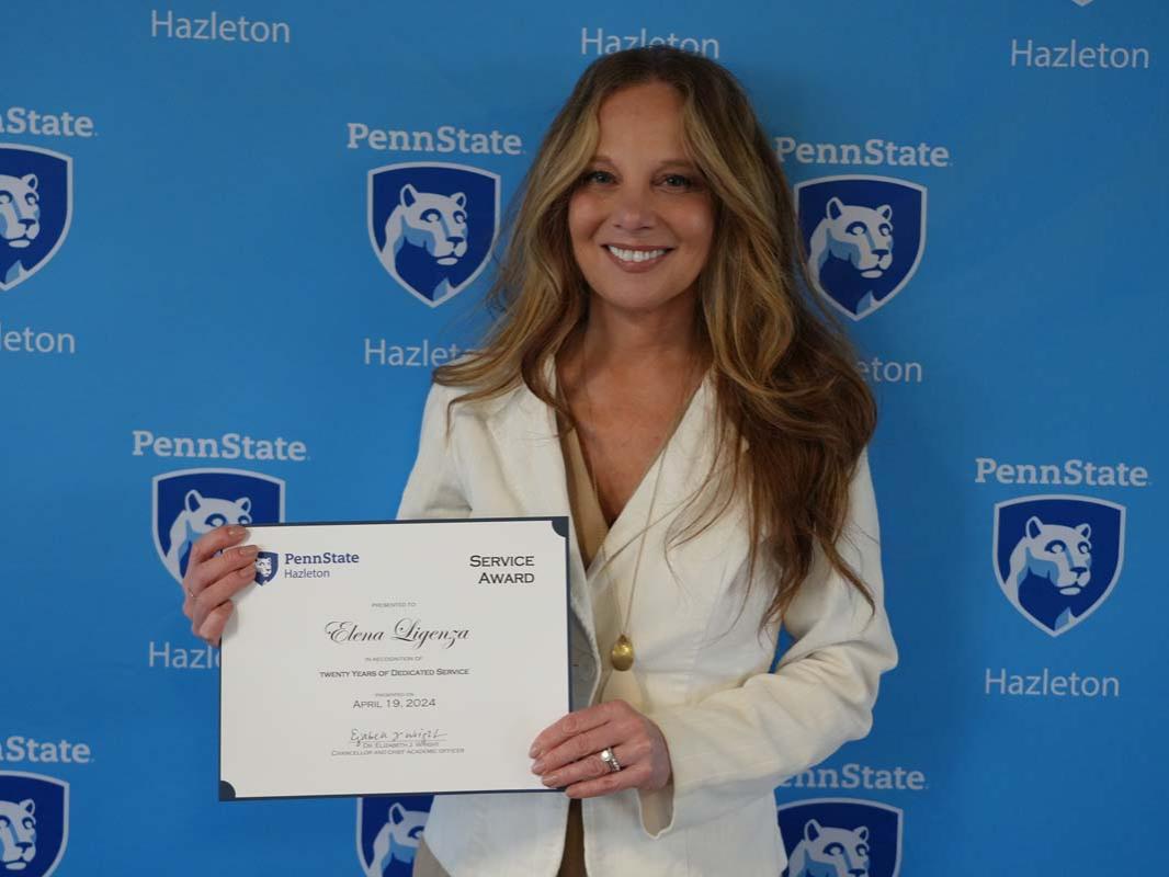 Woman with blonde hair and white jacket standing with certificate in front of light blue Penn State Hazleton backdrop.