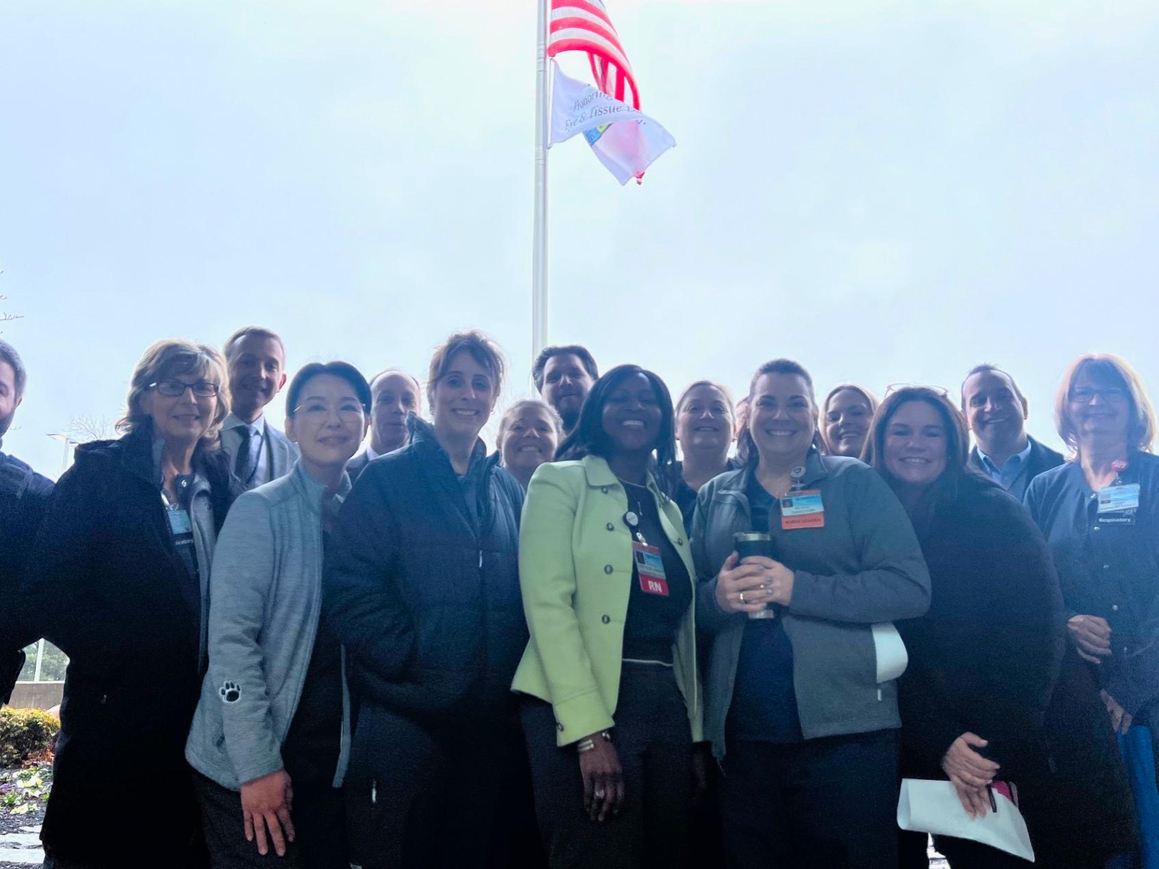 Several people pose for a photo at the base of a flagpole where an American flag flies at the top with a Donate Life flag just below it.