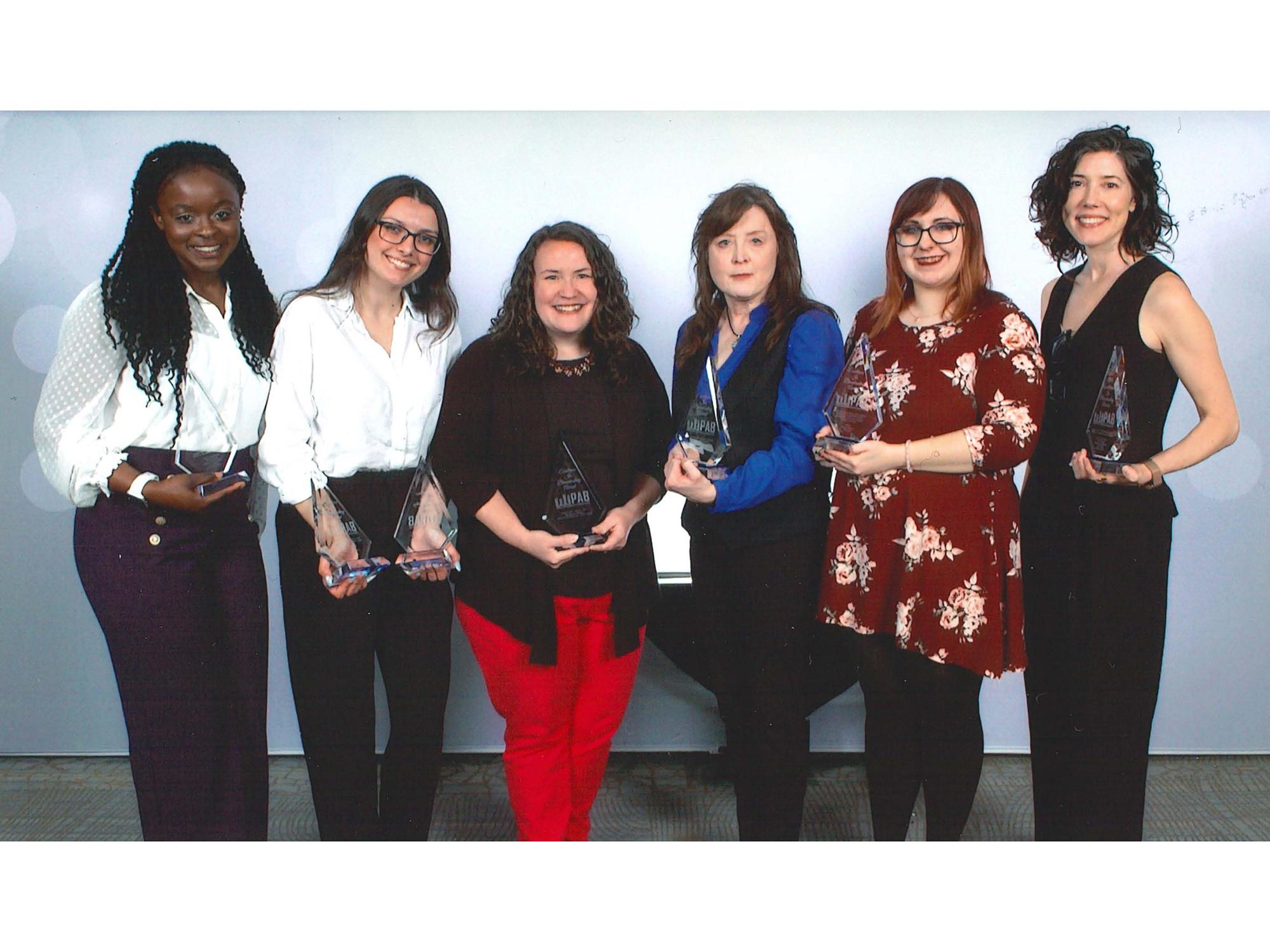 Six women posing in front of blue backdrop, with awards in hand.