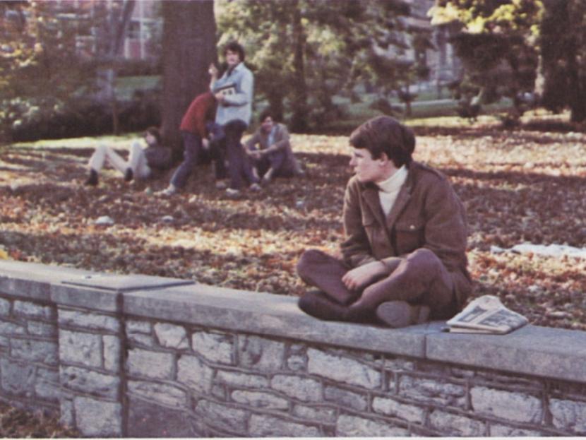 A student sits on a stone wall with a group of students in the background.