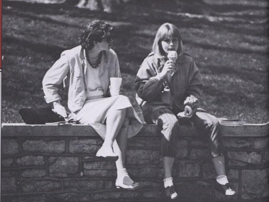 Two students sit on a stone wall. One is eating an ice cream cone.