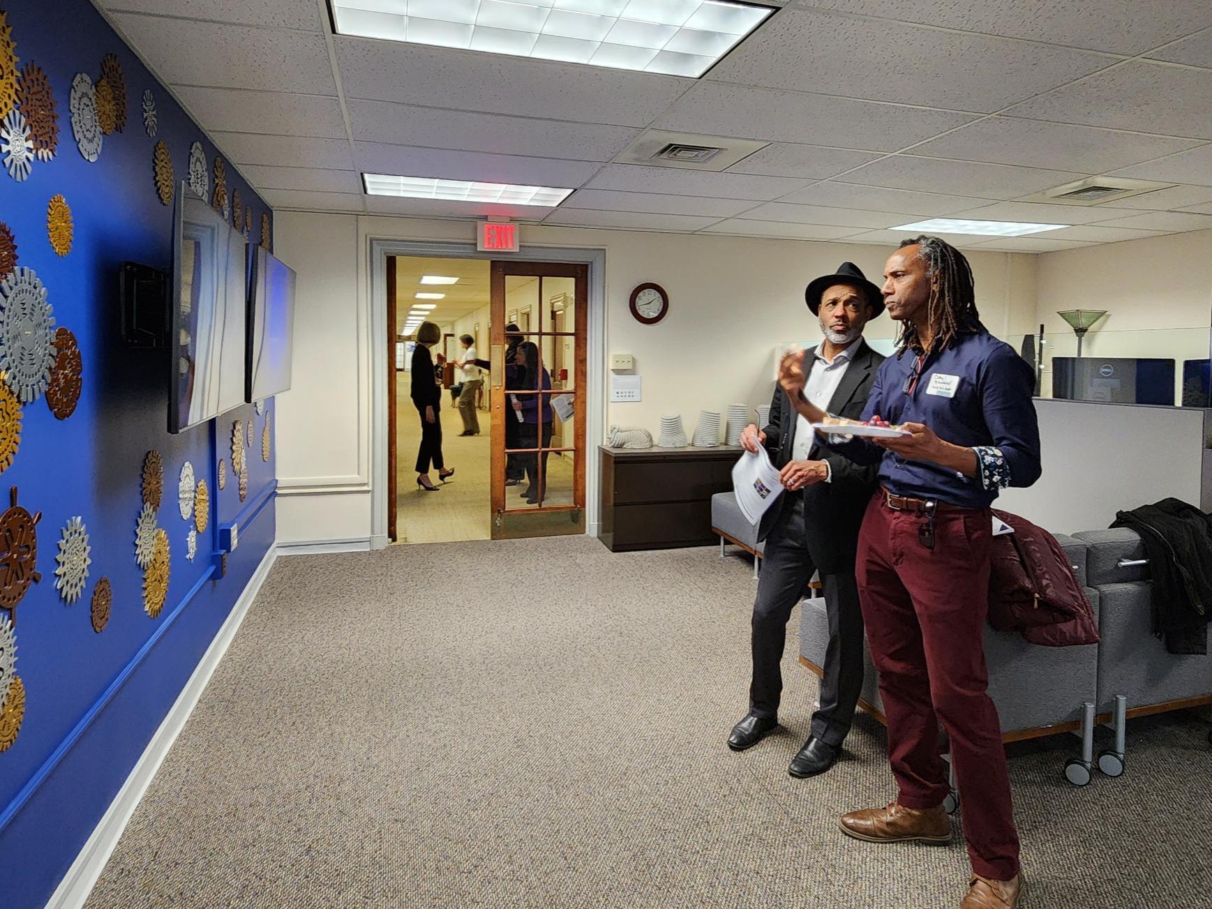 Two people standing in front of a blue wall with metal gears and monitors, talking. 