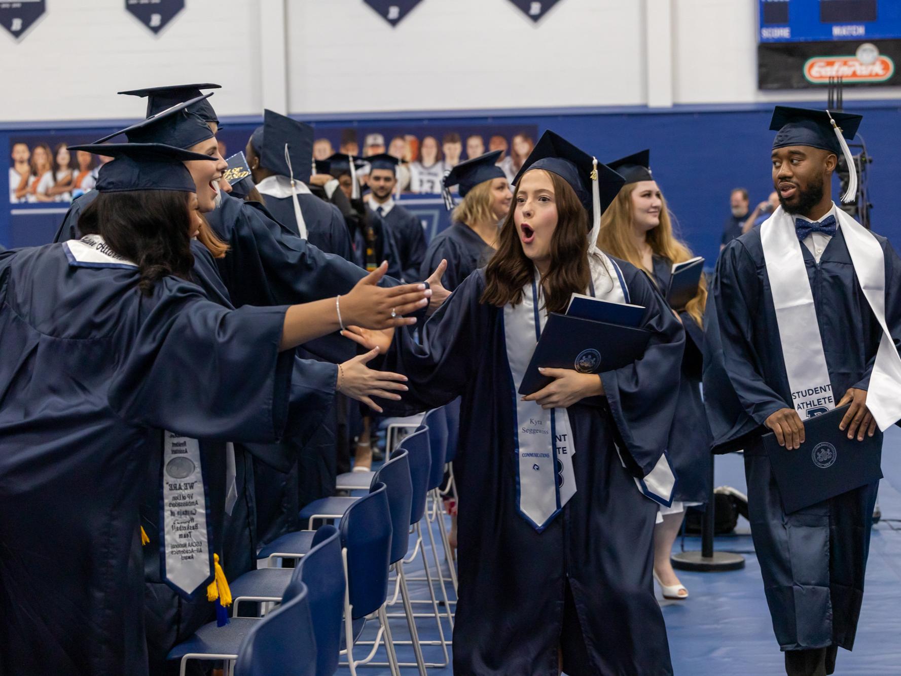 A graduate greets 2 friends as she exits after commencement. All are dressed in caps and gowns.