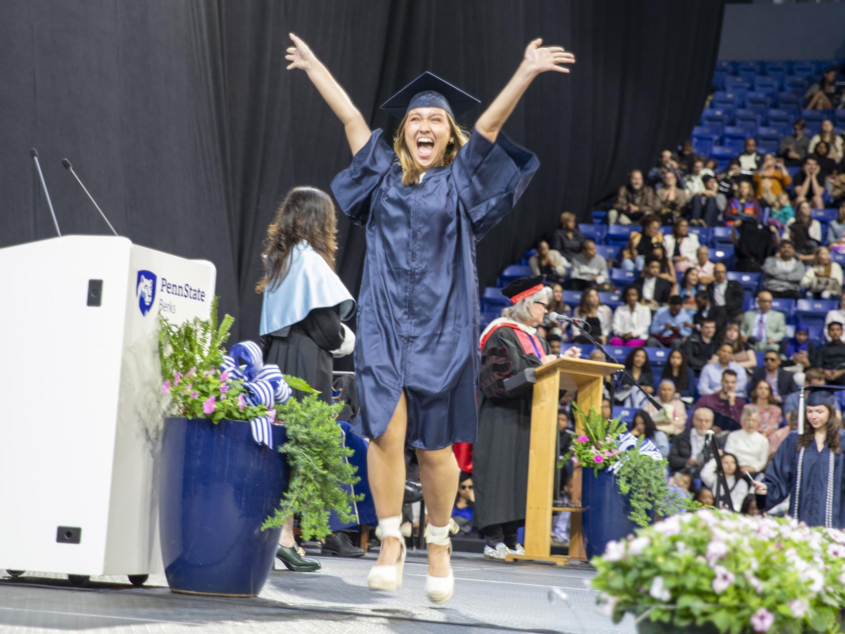 Student wearing cap and gown jumps for joy crossing the stage at commencement