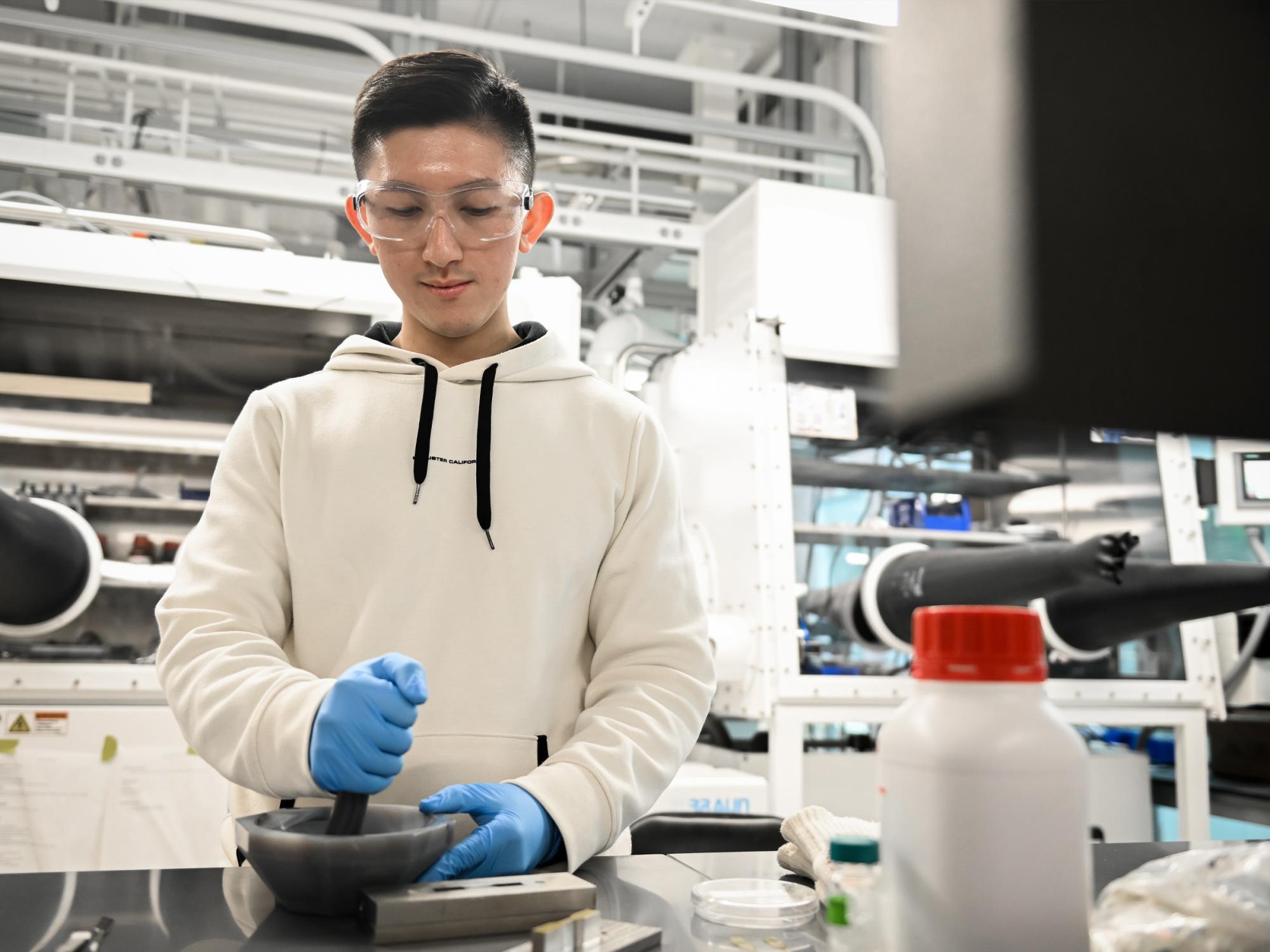 An individual in a lab wearing protective glasses and gloves uses a mixing bowl to combine a sample. 