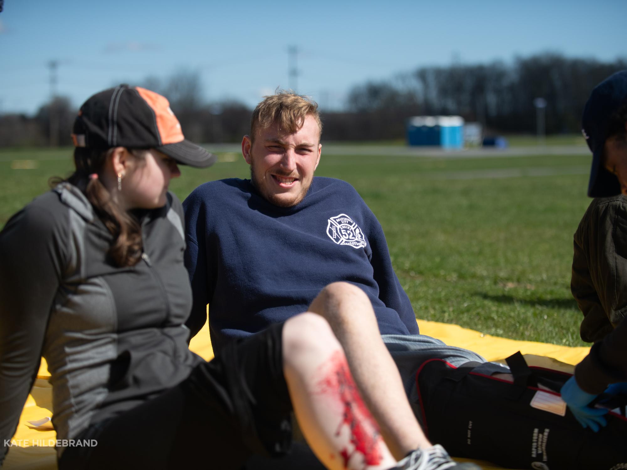 Two students sit on a mat on the ground and are treated for fake leg injuries by another student. 