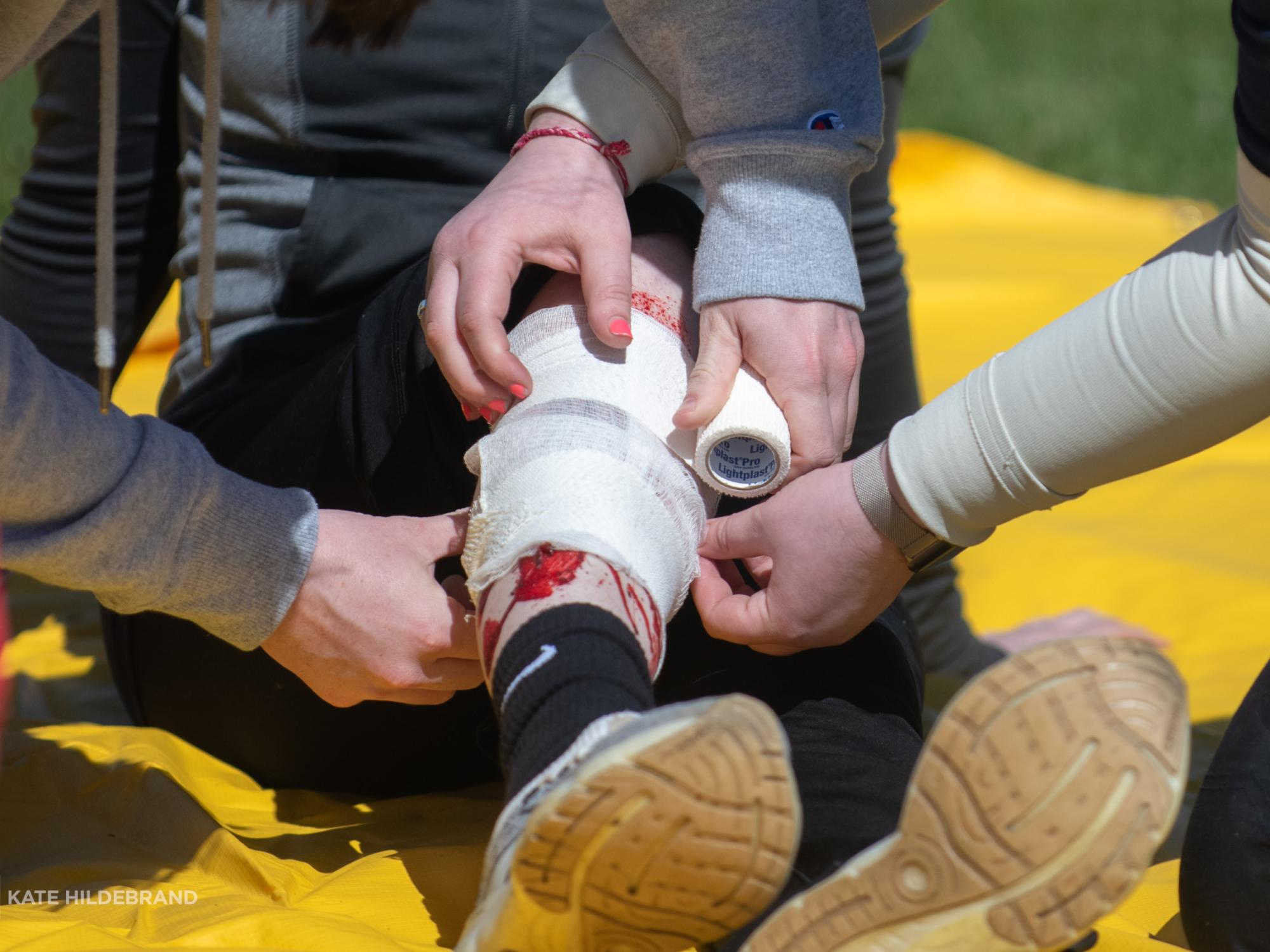 Students wrap gauze around a fake leg wound on another student sitting on the ground.