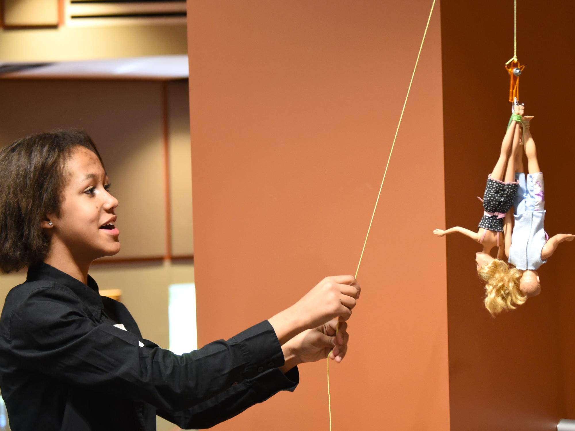 A middle-school student raises a bungee cord that holds two Barbie dolls.
