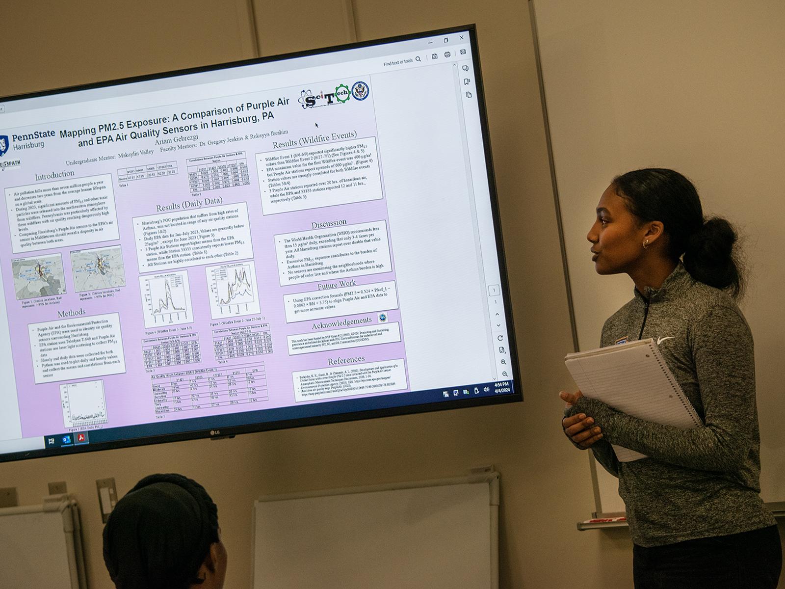 Ariam Gebrezgi stands next to a big screen showing her research project for the EnvironMentors program