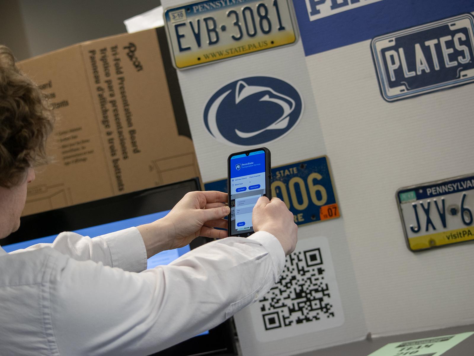 A student holds a phone at arm's length in front of a poster featuring license plates
