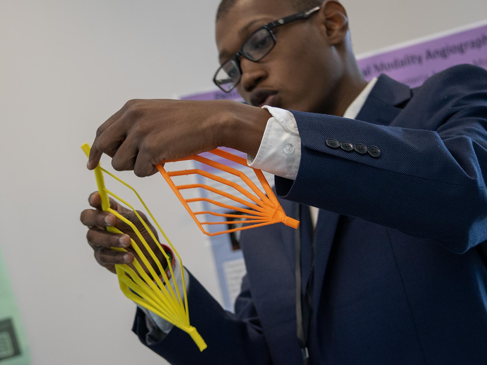 A student in a suit holds two brightly colored objects as he discusses his Capstone project