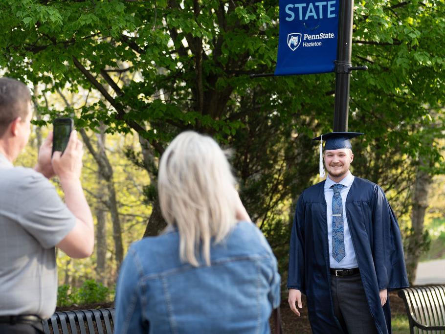 Student in cap and gown having photo taken by man and woman in front of a tree and light post.