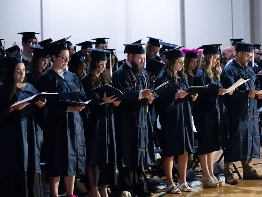 Line of graduates reading from a booklet while standing.