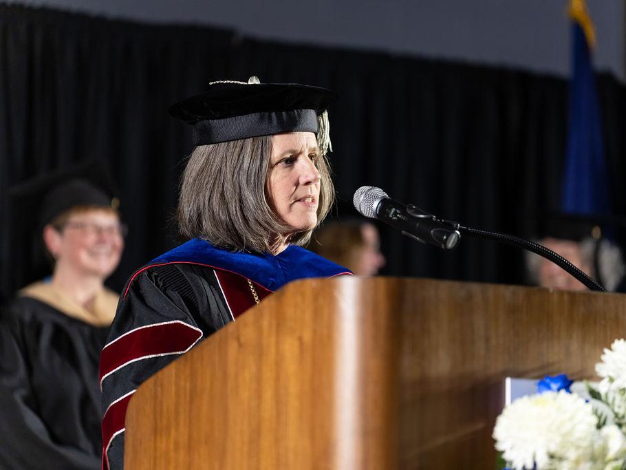 A woman in a dark colored academic regalia delivers remarks at a brown podium.