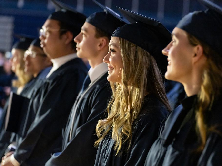 A female graduate smiles and looks toward a stage surrounded in seats by other graduates.