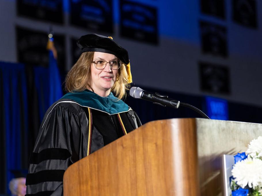 Woman with brown hair in dark academic regalia speaking at a podium.