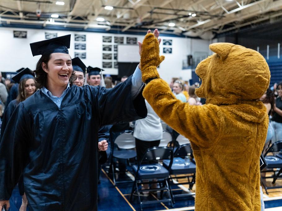 A graduate in a blue cap and gown high fives a Nittany Lion mascot.