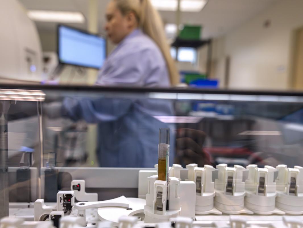 Small, circular containers on a conveyer belt track in a lab. One container holds a test tube. A lab tech works in the background.