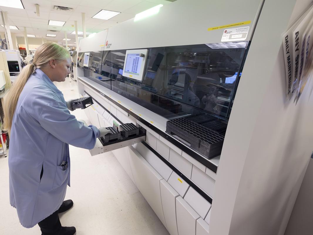 A person in a lab coat stands in front of a long lab testing machine and reaches into a drawer filled with test tubes.