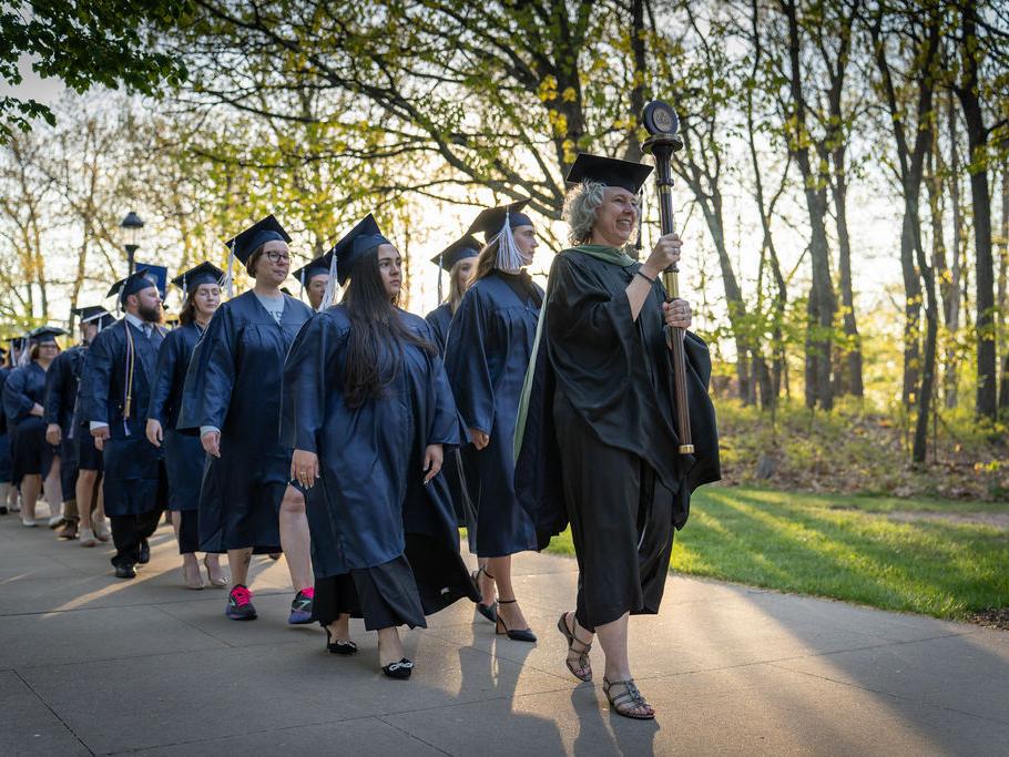 Graduates in caps and gowns being led down a tree-lined sidewalk during a processional.
