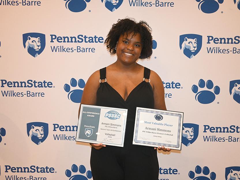A woman holding two certificates standing against a backdrop with the Penn State Wilkes-Barre logo.