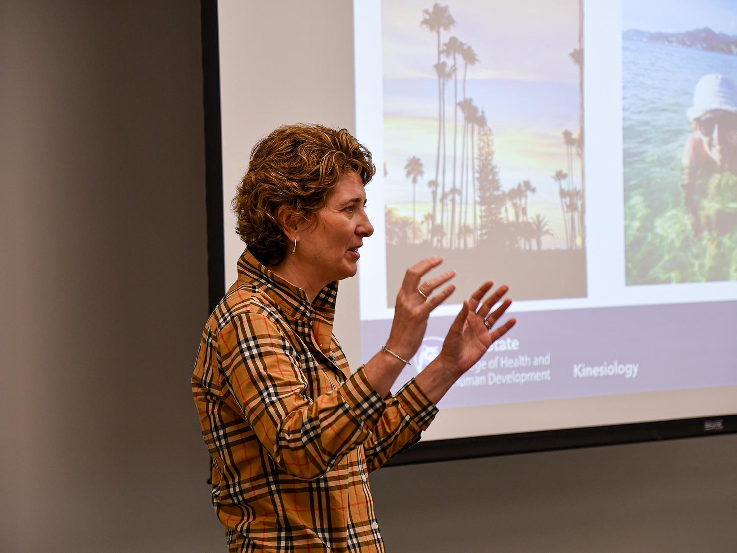 Woman giving a presentation at the front of a room, with large PowerPoint screen