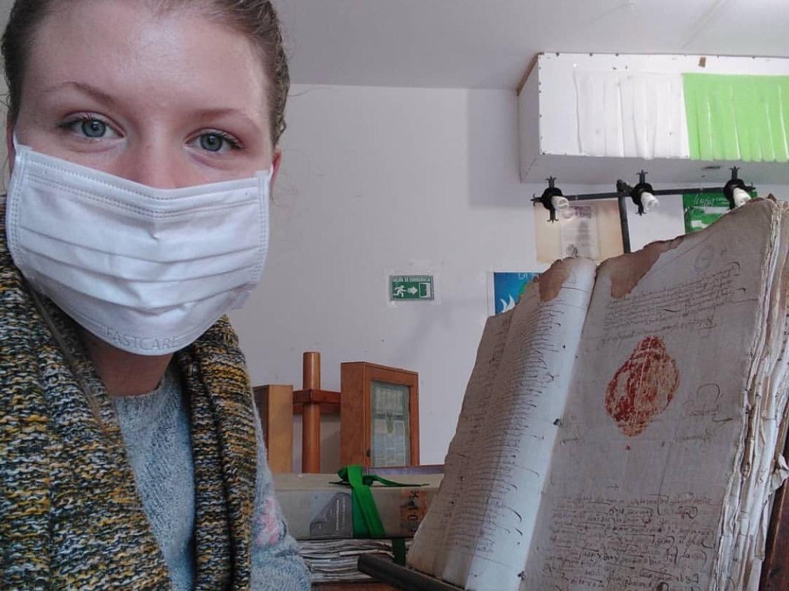 Kate Godfrey wears a mask and sits next to old manuscript 
