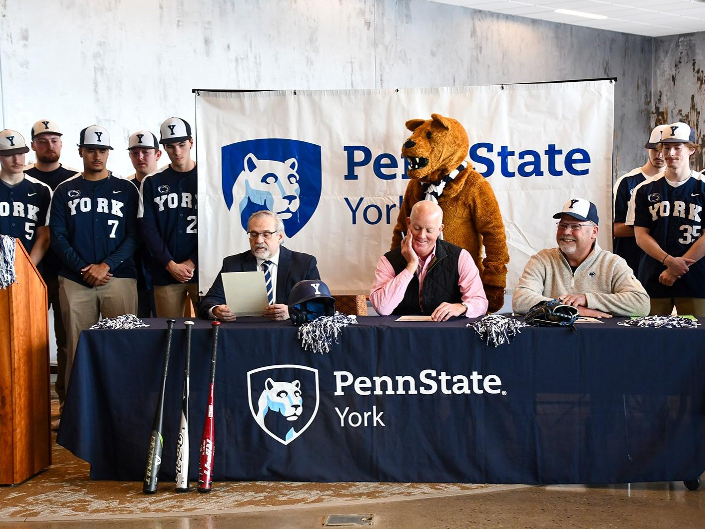 A baseball team stands around three men sitting at a table, with the Nittany Lion mascot behind them.