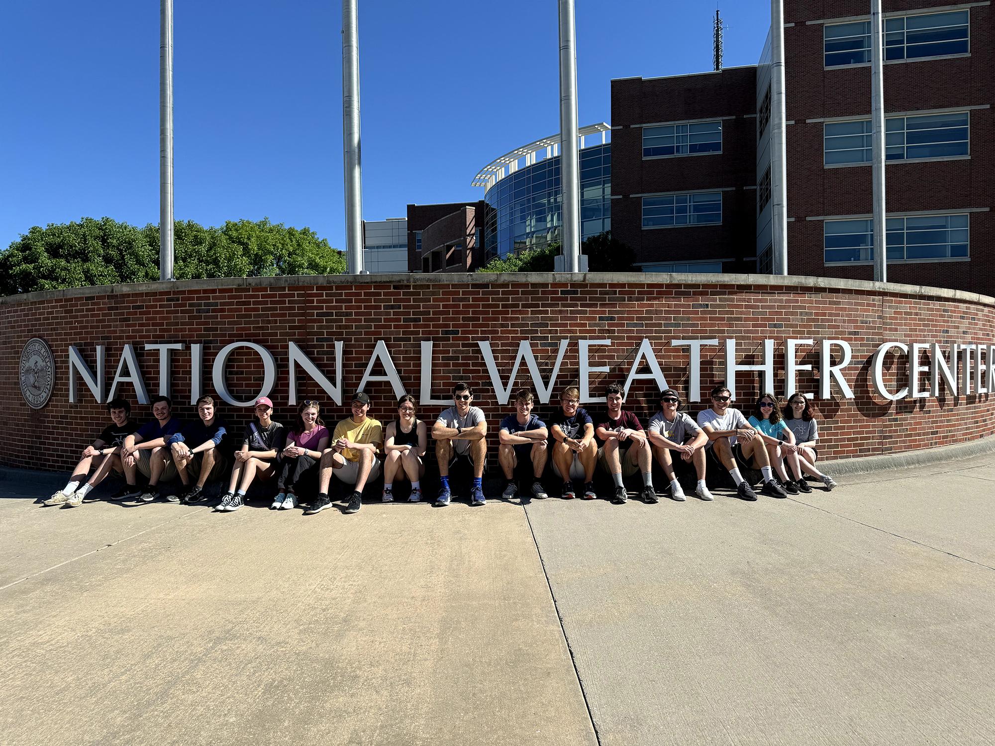 Line of people sitting in front of a brick wall that reads "National Weather Center"