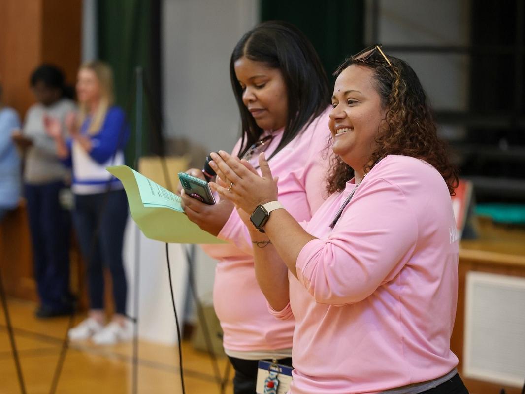 Two people stand in a gymnasium. One is clapping, the other is reading from a piece of paper. Behind them a row of others stand out of focus.