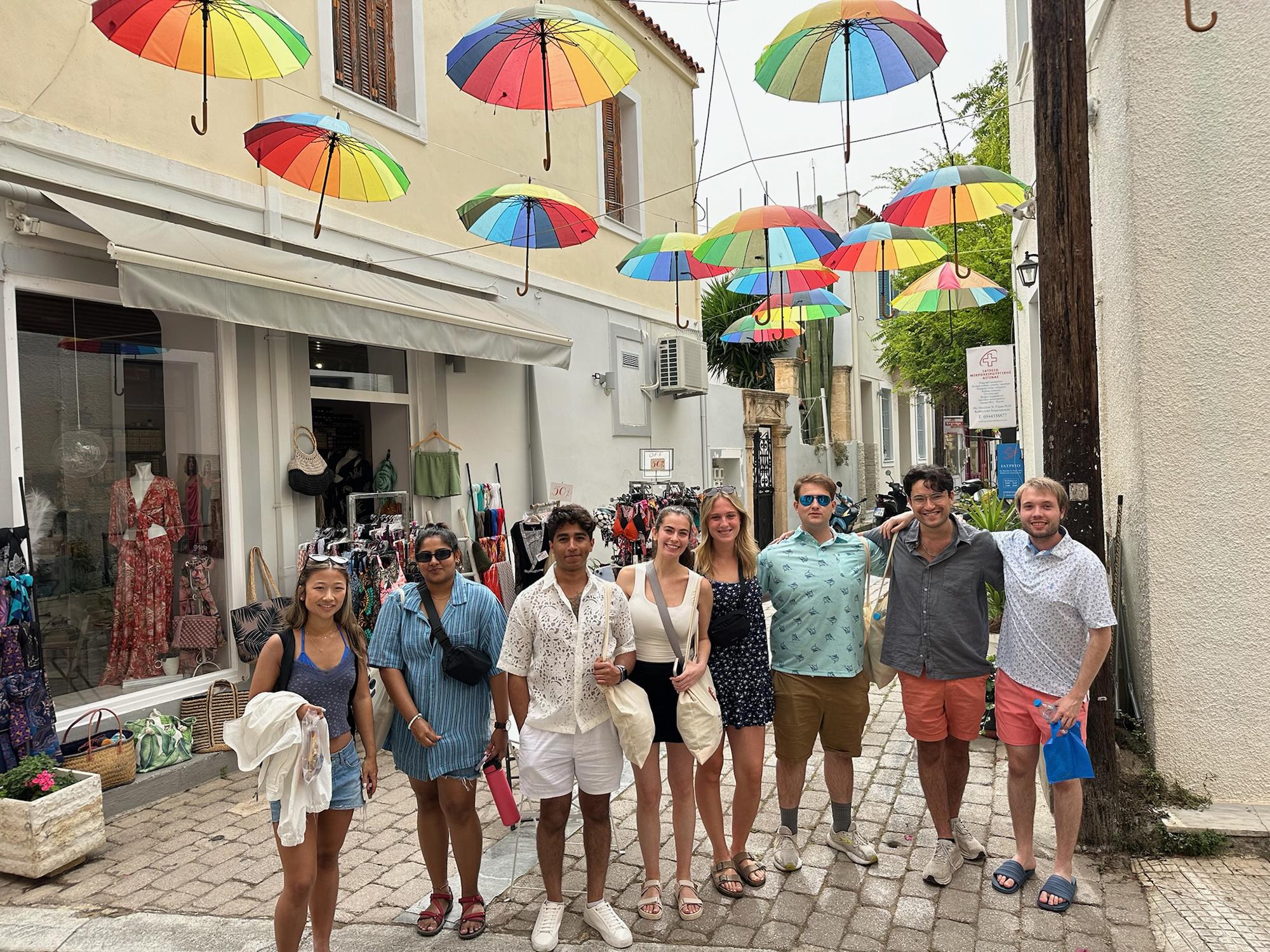 Eight Penn State students stand under rainbow umbrellas on a street in Greece.