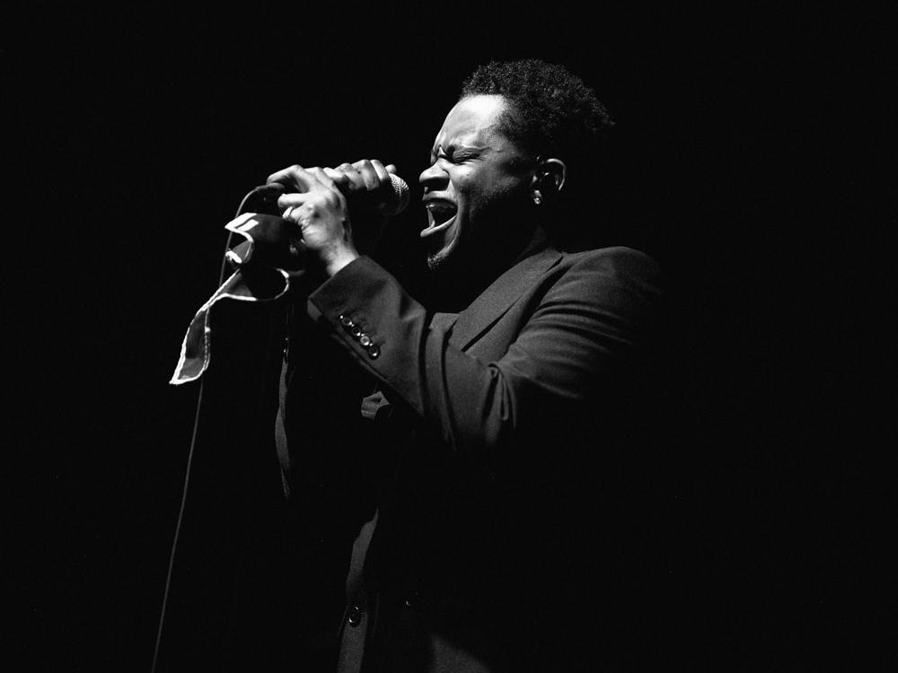 A Black man wearing a dark suit holds a microphone with both hands as he sings into it.