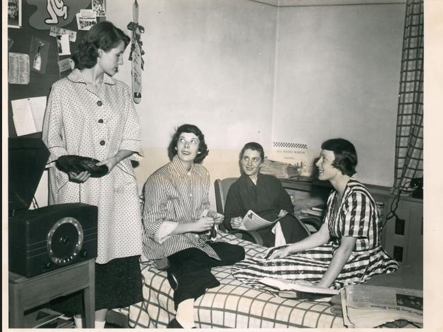 Four students in a dorm room. One holds a record.