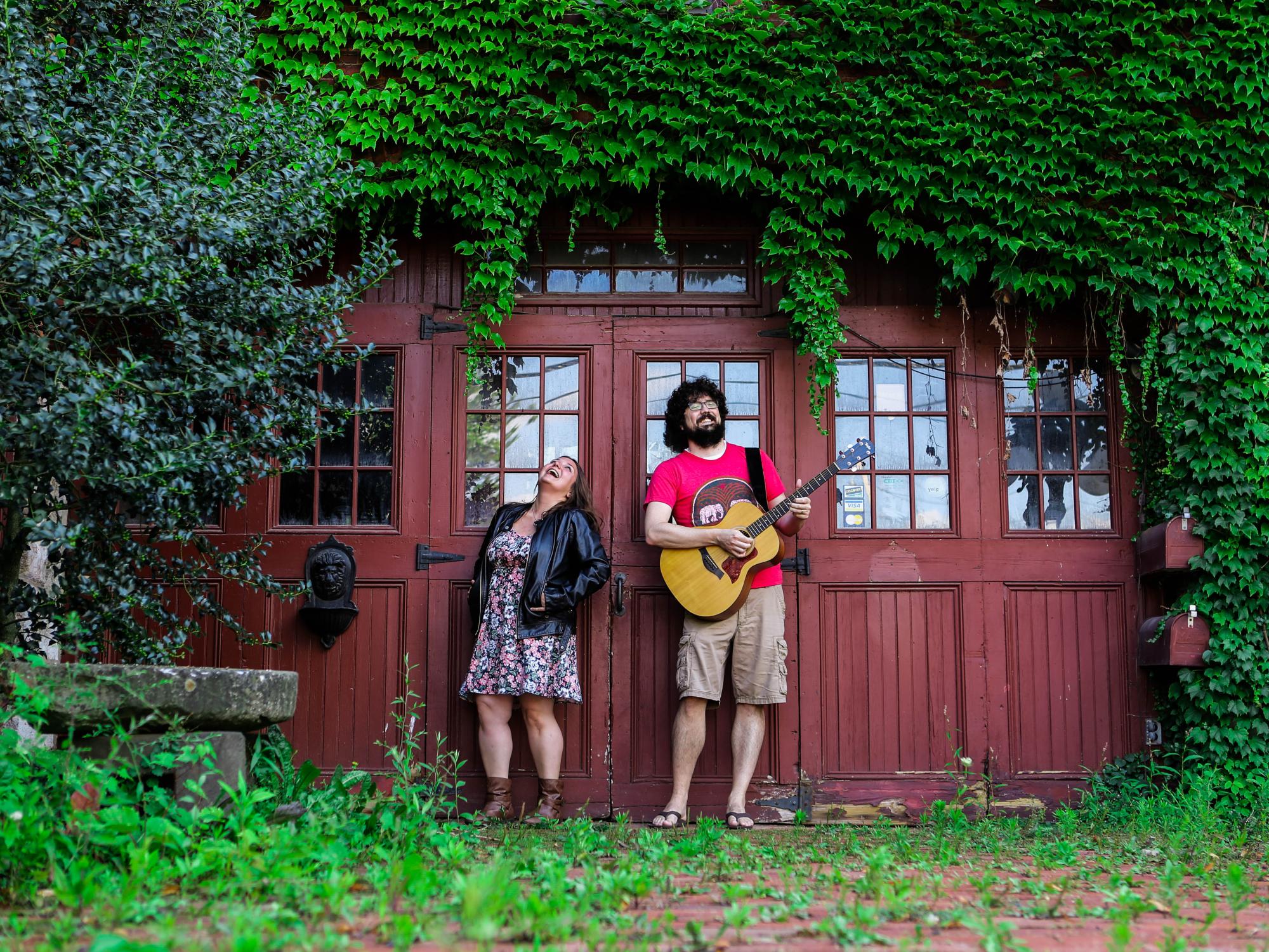 A woman and man holding a guitar stand against an ivy-covered barn door.