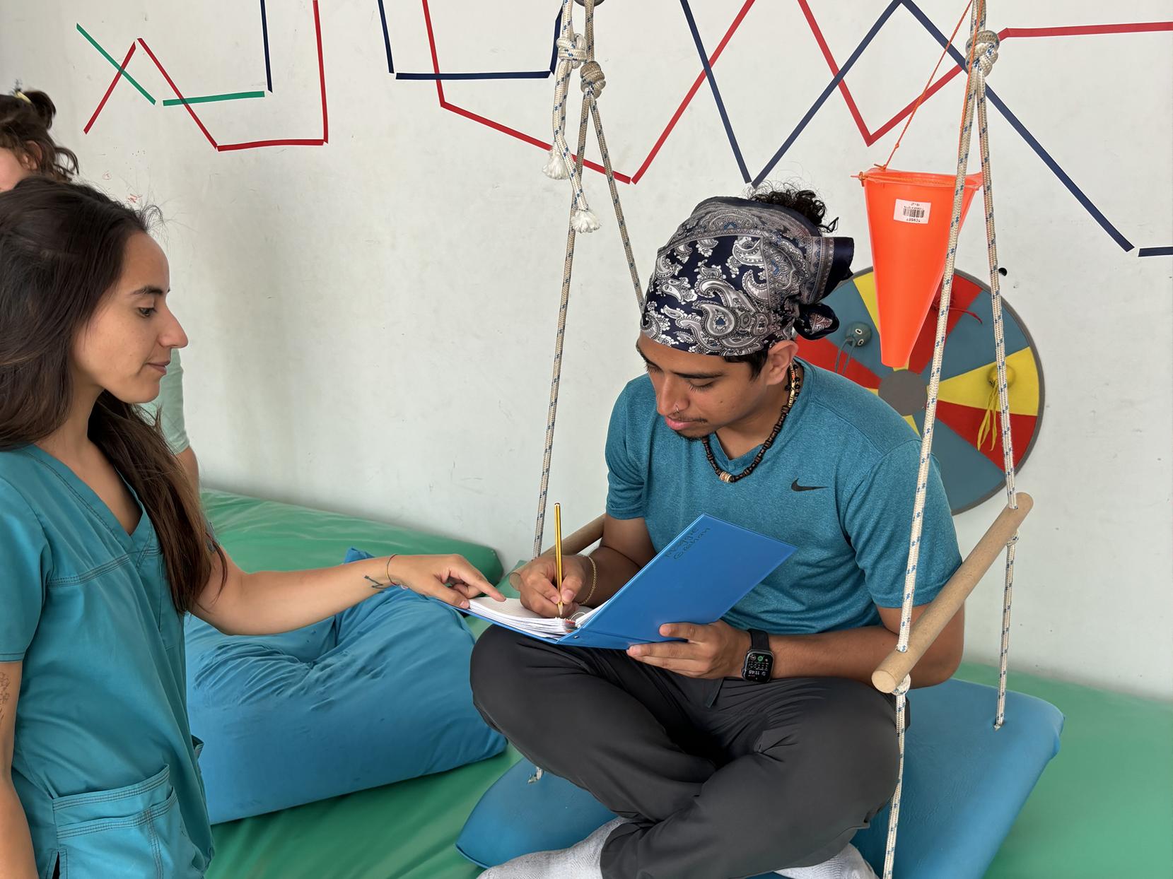 Juan Serrano, working in a therapy room, sits on a piece of equipment and writes in a folder