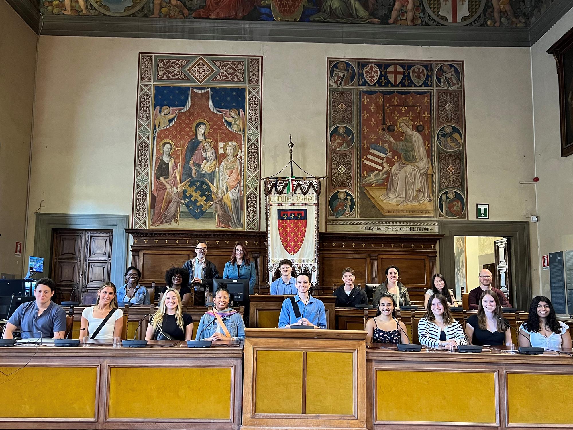 Students and faculty sit in the governmental seats in the Palazzo Comunale of Prato in front of local flags and art.