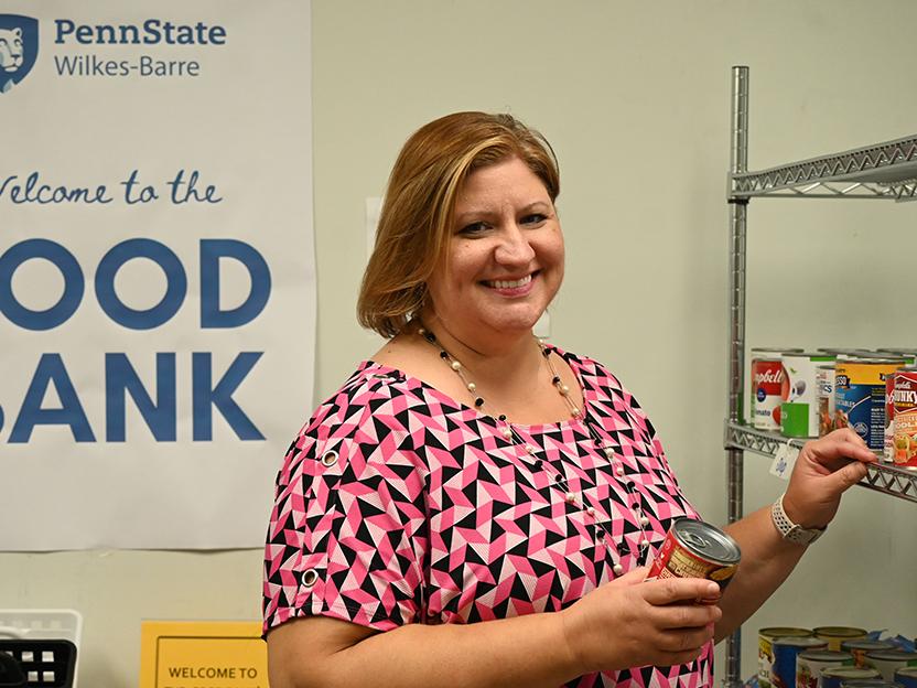 A woman standing next to a rack of canned goods, with the words "food bank" on a sign behind her.