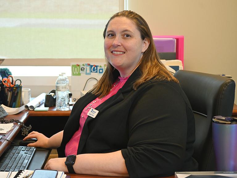 A woman sitting in front of a computer, facing the camera and smiling.