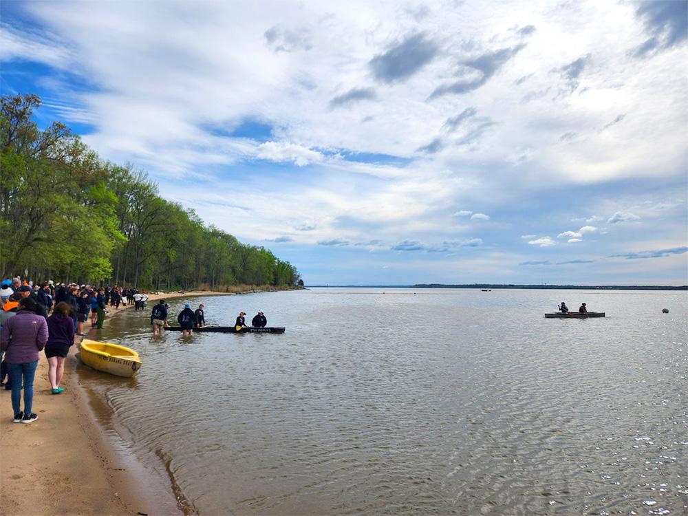  People stand on the shore of a lake while canoes launch from the shore.
