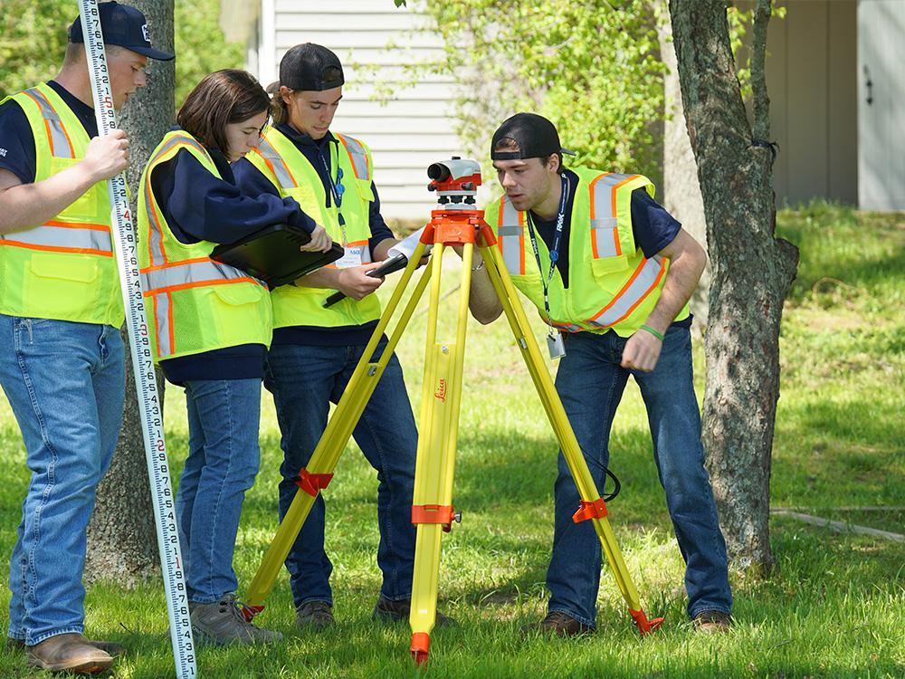 Four people in neon vests consult clipboards and use a piece of surveying equipment.